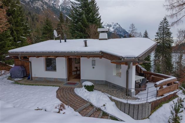 Ein gemütliches Haus im Schnee, umgeben von Bäumen und Bergen. Die verschneite Terrasse lädt zum Entspannen ein.