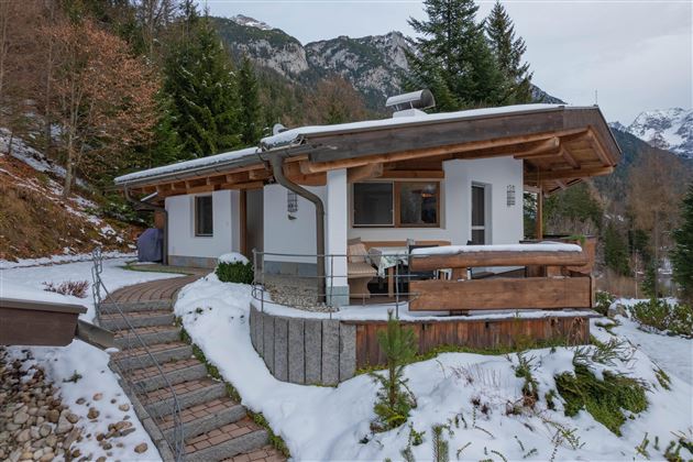 Ein gemütliches Ferienhaus im Schnee, umgeben von Bäumen und Bergen. Der Eingang ist mit einem gepflasterten Weg und einer Holzterrasse gestaltet.