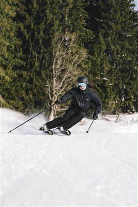 Ein Skifahrer fährt durch den Schnee in einem Wald. Die Umgebung ist winterlich und die Bäume sind grün.