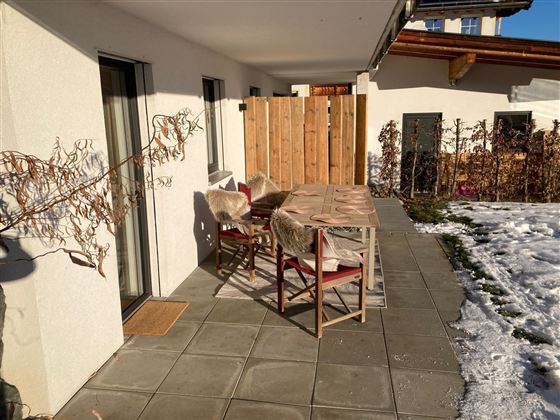 Ein gemütlicher Sitzbereich mit einem Tisch und Stühlen auf einer Terrasse. Im Hintergrund sind Holzverkleidungen und etwas Schnee zu sehen.