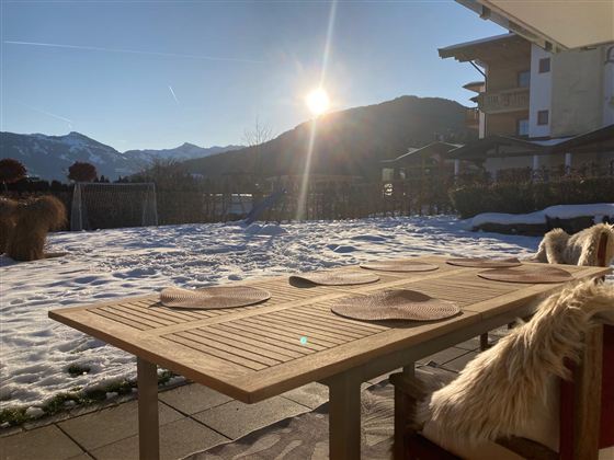 Ein schöner Tisch auf einer Terrasse mit Blick auf die Berge. Die Sonne scheint und der Garten ist mit Schnee bedeckt.