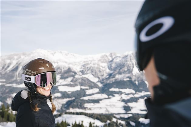 Zwei Skifahrer lächeln sich auf einem schneebedeckten Berg gegenüber. Im Hintergrund sind majestätische Berge und ein klarer Himmel zu sehen.
