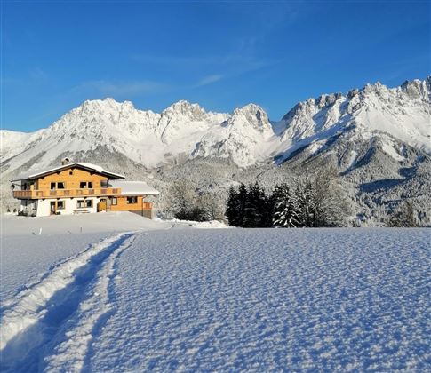 A cozy house is situated in a snow-covered landscape. In the background, majestic mountains rise under a blue sky.