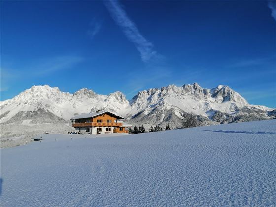 A beautiful wooden house is situated in a snow-covered landscape. In the background, impressive mountain peaks and a clear blue sky can be seen.