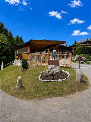A rustic wooden house surrounded by trees and a well-maintained garden. In the foreground, there is a decorative stone with flowers and shrubs.