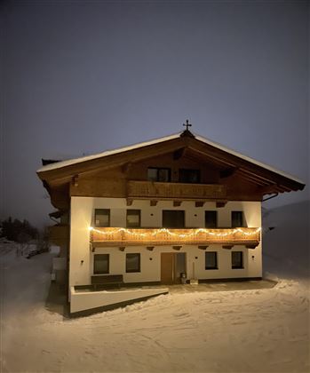 A cozy house in the snow, illuminated by warm lights. The sky is dark and there is a calm winter atmosphere in the air.