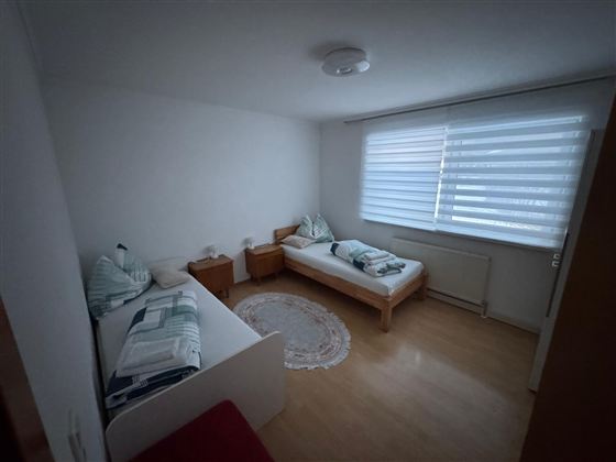 Ein einfaches Zimmer mit zwei Betten und einem runden Teppich. Großer Fenster mit halbtransparenten Jalousien sorgt für sanftes Licht.