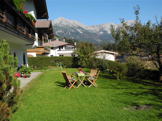 Eine schöne Grünfläche mit zwei Liegestühlen und einem Tisch, umgeben von Bäumen und Blumen. Im Hintergrund sind beeindruckende Berge zu sehen.
