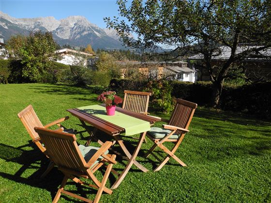 Ein schöner Garten mit einem Tisch und vier Stühlen. Die Berge sind im Hintergrund sichtbar und die Sonne scheint.