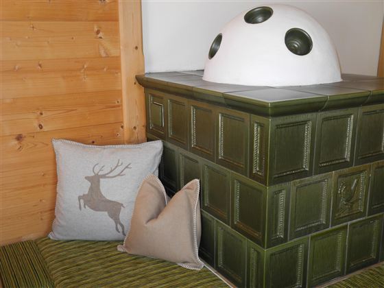 Ein traditioneller Kachelofen mit einem weißen, gewölbten Oberteil. Daneben liegen zwei gemütliche Kissen auf einer Sitzfläche.