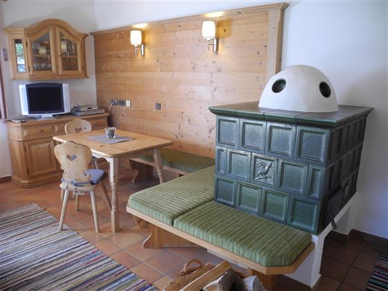 Ein gemütlicher Wohnraum mit Holzverkleidung und einem traditionellen Kachelofen. Ein Essbereich mit einem Tisch und Stühlen ergänzt die warme Atmosphäre.