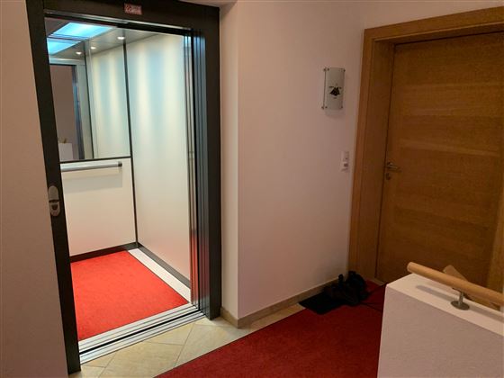 Ein moderner Aufzug in einem hellen Flur mit rotem Teppich. Auf der rechten Seite ist eine Holz- und Glastür sichtbar.