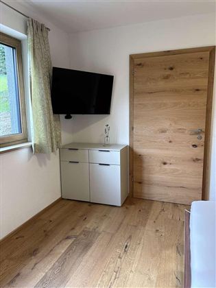 Ein schlichtes Zimmer mit einem Fernseher an der Wand und einem Holztür. Der Boden ist aus Holz und es gibt einen kleinen Schrank unter dem Fenster.