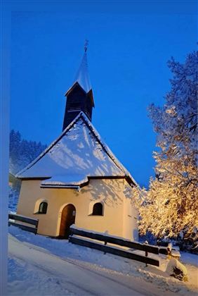 Eine kleine Kapelle im Schnee bei Dämmerlicht. Die Bäume sind mit Schnee bedeckt und die Umgebung strahlt eine ruhige Winteratmosphäre aus.