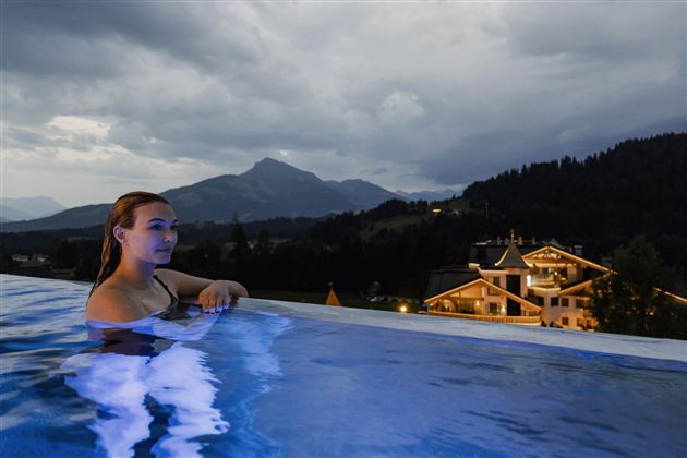 Eine Frau entspannt in einem Infinity-Pool mit Blick auf die Berge. Der Himmel ist bewölkt, und im Hintergrund sind beleuchtete Gebäude zu sehen.