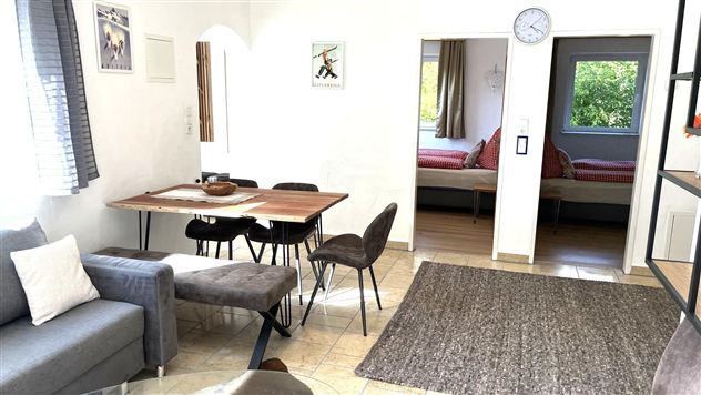 Ein helles Wohnzimmer mit einem Esstisch und gemütlichen Stühlen. Im Hintergrund sind zwei Betten und ein Fenster mit Gartenblick zu sehen.