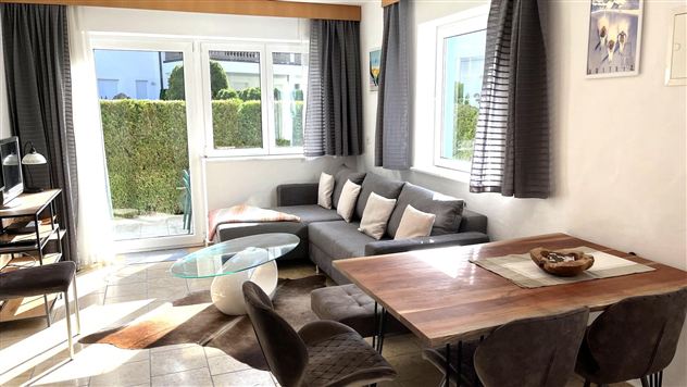 Eine helle und moderne Wohnküche mit einem gemütlichen Sofa und einem Esstisch. Große Fenster lassen viel Tageslicht herein und bieten Blick auf einen gepflegten Garten.