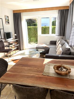 Ein modernes Wohnzimmer mit einem gemütlichen Sofa und einem Holztisch. Große Fenster sorgen für viel Tageslicht und einen Blick ins Grüne.