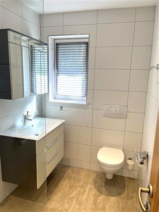 Ein modernes Badezimmer mit großen Fliesen und einem schlichten Design. Es verfügt über ein Waschbecken, einen Spiegel und eine Toilette.