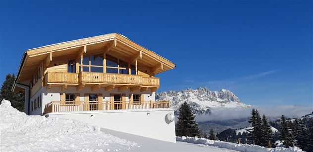A modern chalet in the mountains, surrounded by snow. In the background, majestic mountains and a blue sky can be seen.