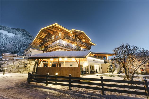 Ein charmantes Berghaus im Schnee, beleuchtet von warmem Licht. Im Hintergrund sind schneebedeckte Berge sichtbar.