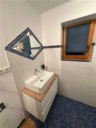 Ein modernes Badezimmer mit einem Waschbecken und einem Spiegel. Die Wände sind hell gefliest, und es gibt ein Fenster mit Jalousien.