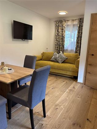 Ein gemütliches Wohnzimmer mit einem gelben Sofa und einem Esstisch. An der Wand hängt ein Fernseher und das Zimmer hat helle Holzfußböden.