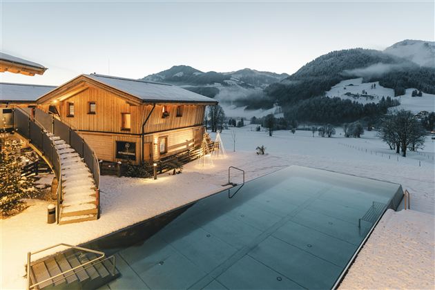 Ein modernes Holzhaus im Schnee mit einer großen Fensterfront. Im Vordergrund befindet sich ein fast leeres Schwimmbecken und im Hintergrund sind sanfte, schneebedeckte Hügel zu sehen.