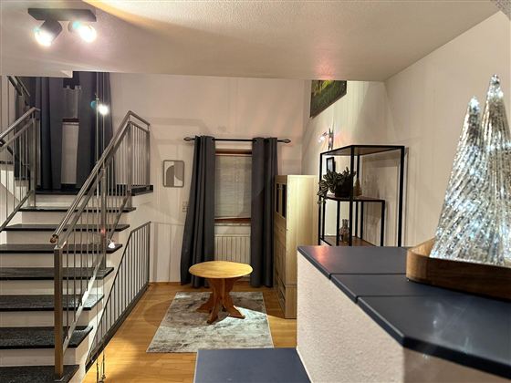 Ein modernes Wohnzimmer mit einer Treppe im Hintergrund. Im Vordergrund steht ein runder Holztisch auf einem Teppich.