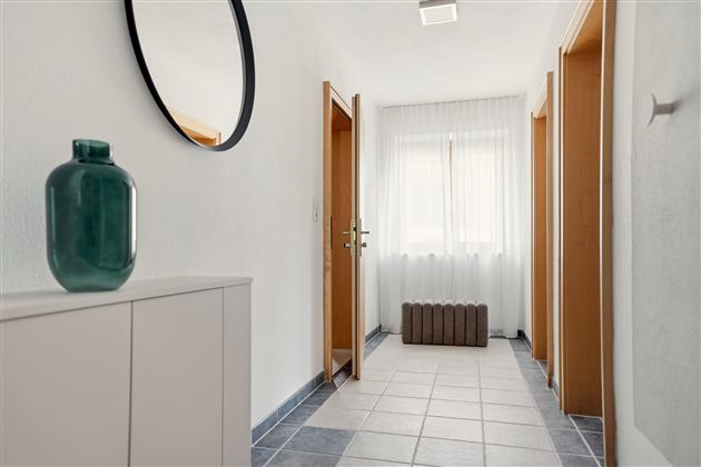 Ein heller Flur mit einem modernen Sideboard und einer Vase. Die Wände sind weiß, und es gibt große Fenster mit Vorhängen.