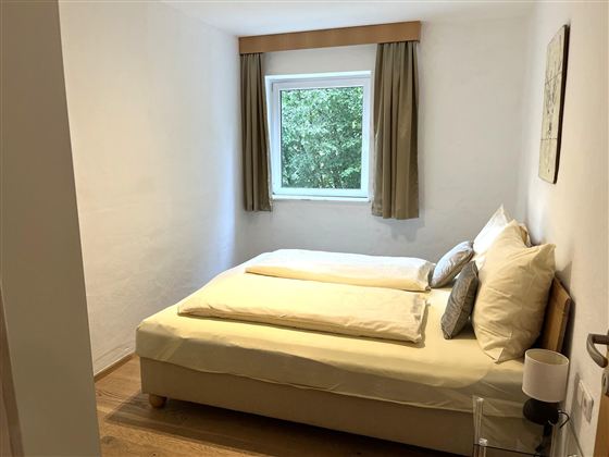 Ein gemütliches Schlafzimmer mit einem Doppelbett und frischen Bettwaren. Das Zimmer hat ein Fenster mit Blick auf Bäume und helle Wände.