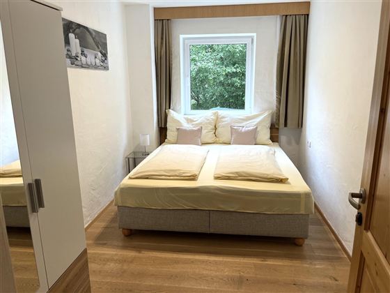 Ein gemütliches Schlafzimmer mit einem großen Bett und hellen Kissen. Fenster mit Blick ins Grüne und moderne Möbel schaffen eine einladende Atmosphäre.