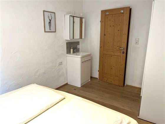 Ein einfaches Schlafzimmer mit einem Doppelbett und einem Holzschrank. Die Wände sind weiß und eine Holz-Tür führt nach außen.