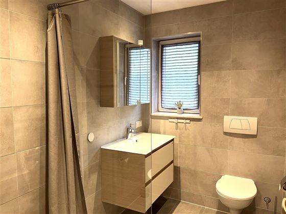 Ein modernes Badezimmer mit hellen Fliesen und einer Dusche. Es gibt ein Waschbecken mit einem Spiegel und ein Fenster mit Jalousien.
