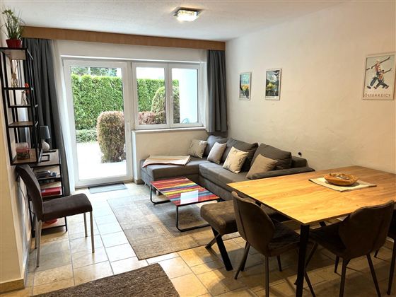 Ein modernes Wohnzimmer mit einer grauen Couch und einem Esstisch. Durch große Fenster scheint Tageslicht und der Blick geht auf einen grünen Garten.