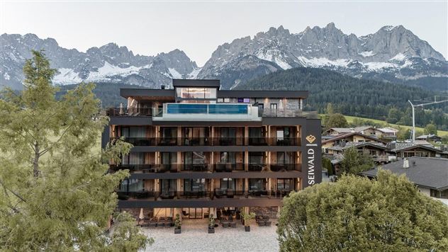 Ein modernes Hotel mit großflächigen Balkonen, umgeben von hohen Bergen. Im Vordergrund befinden sich Bäume und traditionelle Häuser, die eine malerische Landschaft schaffen.