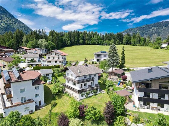 A picturesque landscape with several houses amidst trees and meadows. In the background, gentle hills and a blue sky can be seen.
