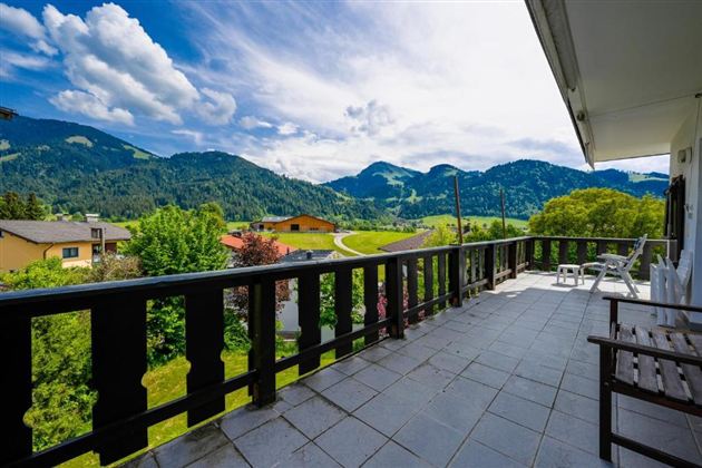 A beautiful balcony overlooking the green mountains and the blue sky. In the foreground, there are chairs, and the surroundings are lush and inviting.
