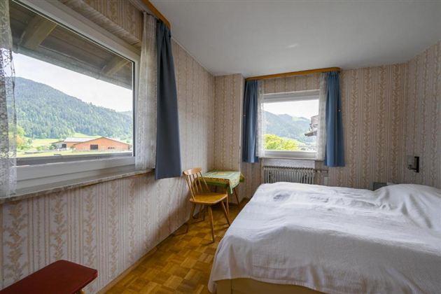 A cozy bedroom with a large bed and a table. Through the windows, there is a beautiful view of the mountains.