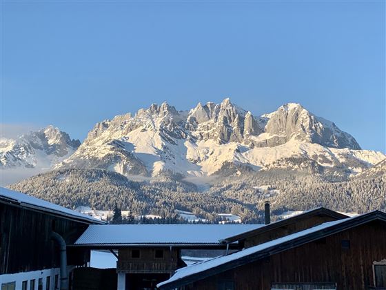 A snow-covered mountain range under a clear blue sky. In the foreground, there are buildings that surround the rural landscape.