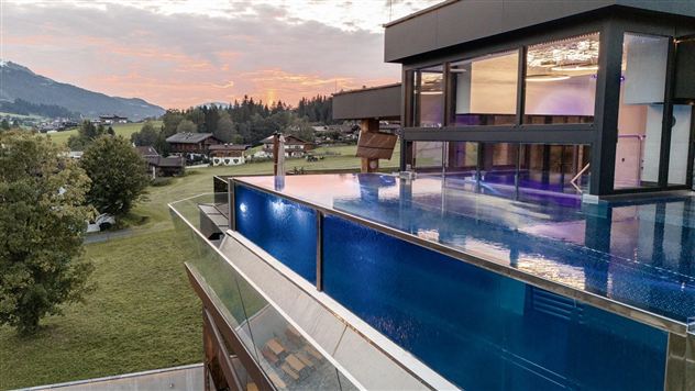 Een modern huis met een infinitypool die in het landschap overgaat. Op de achtergrond zijn zachte heuvels en een kleurrijke zonsondergang te zien.
