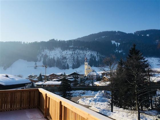 Eine malerische Winterlandschaft mit schneebedeckten Bergen und einem kleinen Dorf. Im Vordergrund ist ein Holzgeländer zu sehen.