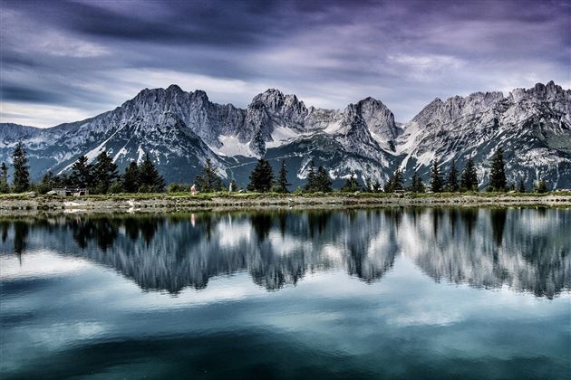 An impressive mountain landscape with snow-capped peaks is reflected in the calm water of a lake. The sky is cloudy, giving the scene a dramatic atmosphere.