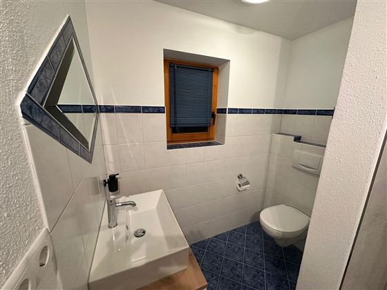 Ein modernes Badezimmer mit weißen Fliesen und blauen Akzenten. Es gibt ein Waschbecken, eine Toilette und ein Fenster mit Jalousien.