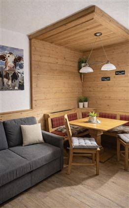 A cozy dining area with wooden paneling and a large table. Next to it is a gray sofa, and two bright lamps hang from the ceiling.