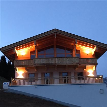 A modern chalet with wooden cladding and a spacious balcony. The lighting creates a cozy atmosphere at dusk.