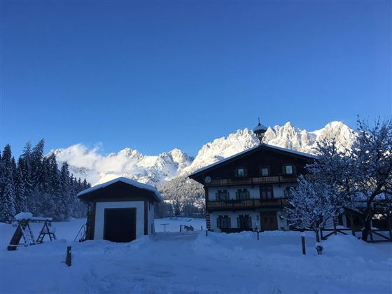 A picturesque winter landscape with snow-covered mountains in the background. In the foreground, there is a traditional alpine cabin and a wooden shed.