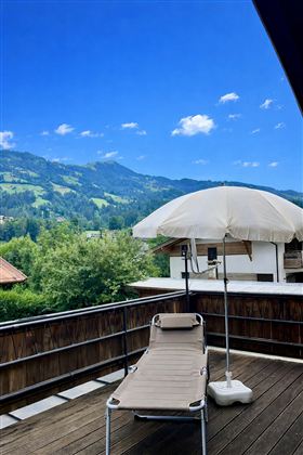 A balcony with a deck chair and a sun umbrella. In the background, green mountains and a blue sky can be seen.