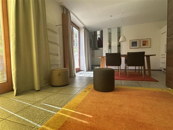 Een lichte woonkamer met een groot raam en houten kozijnen. Een tafel met stoelen staat op een kleurrijk tapijt, en er is een gezellige zithoek.