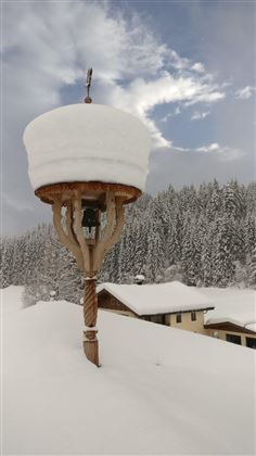 A lantern stands on a snow-covered roof, surrounded by a winter landscape. The trees in the background are also wrapped in snow, while the sky is cloudy.